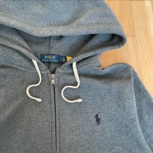 Grå hoodie från Polo Ralph Lauren - Snygg grå hoodie från Polo Ralph Lauren med klassisk logga på bröstet. Tröjan har huva med vita snören, dragkedja framtill och två fickor. Perfekt för chill dagar och enkel att matcha med jeans eller joggers.