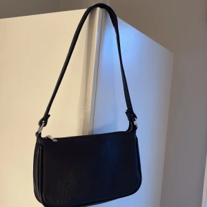 Svart axelväska i skinnimitation - En stilren svart axelväska med minimalistisk design och silverfärgad dragkedja. Väskan har en lång rem som gör den enkel att slänga över axeln och passar perfekt till en trendig vardagslook. Materialet är skinnimitation och formen är avlång och smidig.
