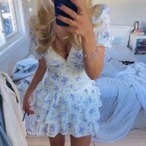 Blommig vit kortklänning med volanger - Supersöt kortklänning i vitt med blått blommönster och volanger. Klänningen har v-ringning, puffiga korta ärmar och dragkedja i sidan. Jättebra skick!