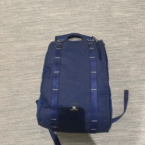 Mörkblå ryggsäck från Db - Säljer en stilren mörkblå ryggsäck från Db med vadderade axelremmar och praktiskt bärhandtag. Väskan har minimalistisk design, svarta detaljer och Db-logga både på rem och ryggpanel. Perfekt för skola, träning.                                                          Nypris 1500
