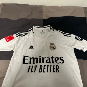 Real Madrid Hemmaställ #5 Bellingham - Säljer en officiell Real Madrid matchtröja med Bellingham och nummer 5 på ryggen. Tröjan är vit med svarta detaljer, Adidas-logga, klubbmärke och sponsortryck. La Liga-märke på ärmen. Tillverkad i lätt funktionsmaterial, perfekt för fotboll eller häng. Storlek S men passar även M, använd nån gång, inga defekter. Priset kan diskuteras.