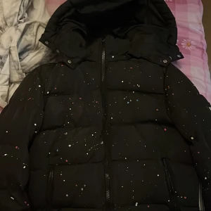 Svart pufferjacka från NVLTY med färgstänk - Säljer en svart pufferjacka från NVLTY med coola färgstänk i vitt, blått och rött. Jackan har huva, dragkedja framtill och två fickor med dragkedja. Perfekt för dig som vill sticka ut med en unik look under kalla dagar.