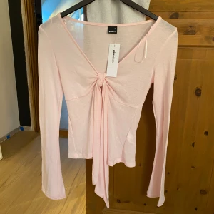 Ljusrosa långärmad topp från Gina Tricot - Supersöt ljusrosa långärmad topp från Gina Tricot med knyt framtill och v-ringad hals. Tunn och lätt i materialet, perfekt för lager-på-lager eller som statement-piece. Snyggt croppad passform och långa ärmar ger en trendig vibe. Helt ny med lapp på och har aldrig använt!💕 