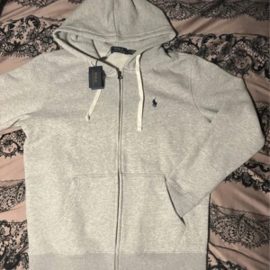 Grå hoodie från Polo Ralph Lauren - Snygg grå hoodie från Polo Ralph Lauren med klassisk broderad logga på bröstet. Tröjan har huva med vita snören, dragkedja framtill och två stora fickor. Perfekt för chill dagar och enkel att matcha med jeans eller joggers.