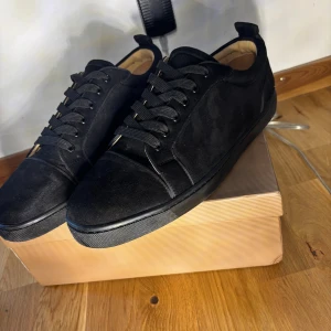 Svarta sneakers från Christian Louboutin - Snygga svarta sneakers från Christian Louboutin i mocka med klassisk röd sula och broderad logga på hälen. Skorna har rund tå, svarta skosnören och diskret design som ändå sticker ut med den ikoniska röda undersidan.