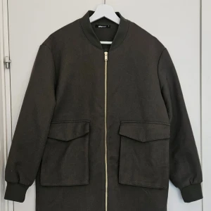 Mörkgrön jacka  - Stilren och minimalistisk jacka från Gina Tricot i mörkgrön färg med guldig dragkedja och stora fickor framtill. Modellen har rak passform med ribbade muddar vid ärmslut och hals.               Använd få gånger, Väldigt fint skick. Strl M