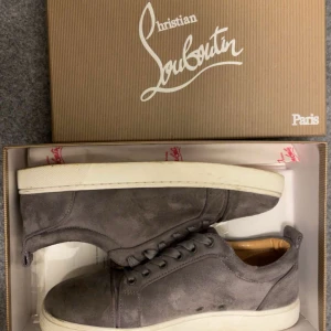 Christian Louboutin skor - Snygga grå sneakers från Christian Louboutin i mjuk mocka med klassisk röd sula. Skorna har rund tå, platt vit sula och ton-i-ton snörning. Insidan är fodrad i beige skinn för extra komfort. Perfekta för dig som vill ha en lyxig och stilren look.