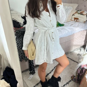 Vit broderad klänning med knyt - Superfin vit kort skjortklänning med broderade blommor, knappar framtill och knytskärp i midjan. Långärmad och i luftig bomull, perfekt för varma dagar. Snygg krage och detaljerade broderier ger en romantisk vibe.