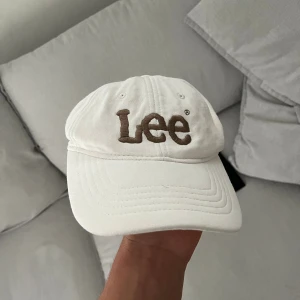 Vit keps från Lee med broderad logga - Snygg vit keps från Lee med stor broderad logga i brunt framtill. Klassisk böjd skärm och justerbar passform bak. Perfekt accessoar för en avslappnad och trendig look.