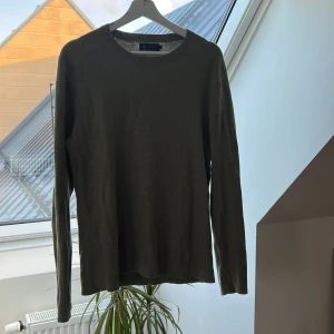 Olivgrön stickad tröja från Riley - En stilren olivgrön stickad tröja från Riley med rund halsringning och långa ärmar. Perfekt för lager-på-lager och enkel att matcha med jeans eller chinos. Tröjan har en klassisk passform och är supermjuk mot huden.