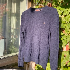 Mörkblå kabelstickad tröja Morris - Kabelstickad mörkblå tröja från Morris i 100% merinoull. Klassisk rund halsringning och diskret broderad logga på bröstet. Perfekt för dig som gillar stilrena och tidlösa plagg med snygg struktur. Sitter skönt och passar till det mesta.