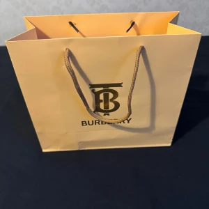 Burberry shoppingväska med klassiskt mönster - Säljer en beige shoppingväska från Burberry med det ikoniska rutig mönstret och en snygg TB-logga i metall. Väskan har rejäla handtag i rep och är tillverkad i slitstark bomull. Perfekt för dig som gillar lyxiga detaljer och klassisk design.