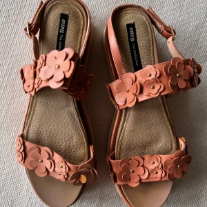 Blommiga platåsandaler från mtng - Snygga sandaler i korallrosa med platåsula och dekorativa blommor på remmarna. Skorna har spänne vid ankeln och mjuk innersula i skinn. Perfekta för att ge din outfit en färgklick och lite extra höjd.
