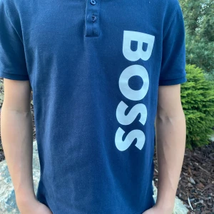 Marinblå BOSS pikétröja  - Snygg marinblå pikétröja från BOSS med stor vit logga längs sidan. Klassisk krage, tre knappar och korta ärmar. Tillverkad i mjuk bomull som känns skön mot huden. Perfekt för dig som gillar stilrena och sportiga plagg. Priset ej hugget i sten. Hör av er vid frågor eller funderingar. 