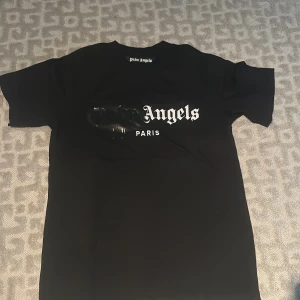 Svart Palm Angels t-shirt Paris  - Svart t-shirt från Palm Angels med Paris-tryck på bröstet och coolt spray-effekt över loggan. Klassisk rund hals och korta ärmar. Materialet är mjuk bomull och passformen är normal. Perfekt för dig som gillar streetwear och snygga prints.