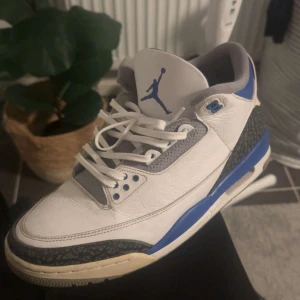 Nike Air Jordan 3 Retro vit/blå - Nike Air Jordan 3 Retro i vitt skinn med blå och grå detaljer. Skon har klassisk snörning, rund tå och ikonisk Jumpman-logga på plösen. Grått mönster runt tån och hälen samt blå accenter på sulan och öglorna. Perfekt för dig som gillar streetwear och sneakerskultur.