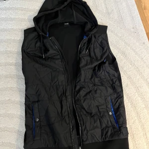 Svart dunväst med huva och blå detaljer (Hugo Boss) - Köpt på Hugo boss i Spanien. Väldigt unik väst. Har använts max 2 gånger och har efter bara legat i garderoben. 