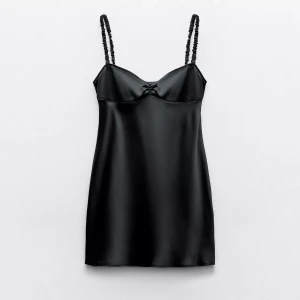 Svart satin kortklänning - Snygg svart kortklänning i satin med smala axelband och liten rosett framtill. Klänningen har en slät, glansig yta och en feminin siluett som ger en chic vibe. Perfekt för dig som gillar stilrena och trendiga plagg med en touch av elegans.