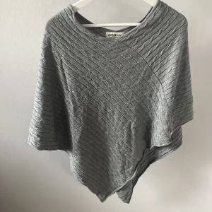 Grå stickad ponchotröja Hampton Republic - Säljer en grå stickad ponchotröja från Hampton Republic. Tröjan har ett diagonalt kabelstickat mönster och en avslappnad, asymmetrisk passform. Perfekt för lager-på-lager och chill dagar. Superenkel att slänga på över en t-shirt eller skjorta.❤️❤️