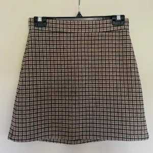 Rutig kortkjol från & Other Stories - Snygg rutig kortkjol från & Other Stories i beige och svart. Kjolen har en klassisk A-linjeform och dragkedja bak. Perfekt för dig som gillar preppy stil och vill ha något som sticker ut lite extra. Materialet känns mjukt och har en lätt struktur.