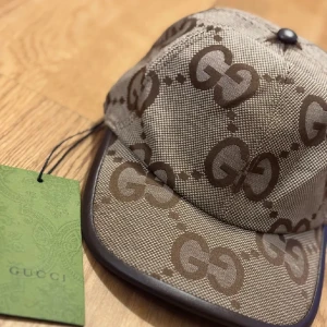 Gucci keps med GG-mönster - Snygg keps från Gucci i beige och brunt med det klassiska GG-logomönstret över hela kepsen. Kepsen har böjd skärm och detaljer i mörkbrunt runt kanten. Tillverkad i Italien och har en exklusiv känsla med märkets gröna tagg kvar. INGÅR ÄVEN BOX