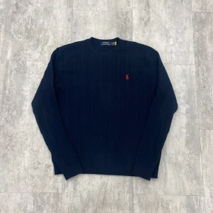 Mörkblå stickad tröja Polo Ralph Lauren - Kabelstickad mörkblå tröja från Polo Ralph Lauren med klassisk röd broderad logga på bröstet. Tröjan har rund halsringning, ribbade muddar och lång ärm. Perfekt för dig som gillar stilren och tidlös design.