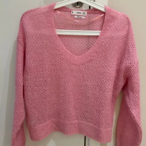Rosa stickad tröja från MNG - Säljer en ljusrosa stickad tröja från MNG i ullblandning. Tröjan har v-ringning, lång ärm och en luftig, lite transparent struktur. Perfekt för lager-på-lager och ger en söt och trendig look.