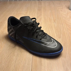 Nike fotbollsskor svart/blå strl 40.5 - Nike Mercurial Vapor 15 Club Ic 'Shadow Pack'. Säljer ett par svarta Nike fotbollsskor med blå sula och vita detaljer. Skorna har snörning och är tillverkade i syntetmaterial med låg profil och platt sula, perfekt för inomhusspel. Snygg design med stor Nike-logga på sidan och tå. Väldigt lite använda. 