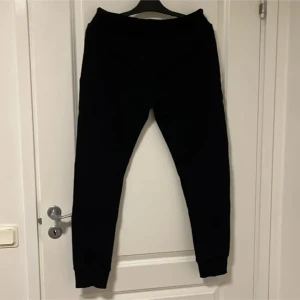 Svarta mjukisbyxor från MR Originals - Svarta mjukisbyxor från MR Originals med smal passform, resår och snörning i midjan samt dragkedjor på fickorna. Byxorna har en liten logga med krona på vänster sida och är tillverkade i mjukt material för maximal komfort.