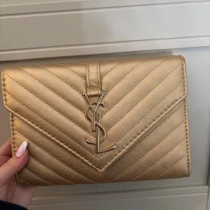 Guldig kuvertväska - Snygg kuvertväska i guldigt skinn med quiltad design och ikoniska YSL-loggan framtill. Väskan har flera fack inuti, inklusive ett med dragkedja, och stängs med magnetlås. Perfekt för dig som vill sticka ut med en lyxig accessoar.