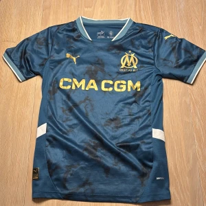Blå Marseille fotbollströja Puma S - Snygg blå Olympique de Marseille fotbollströja från Puma med gul logga och sponsortryck. Tröjan har korta ärmar, svarta mönsterdetaljer och vita inslag vid ärmslut och sidor. Tillverkad i lätt polyester med DryCell-teknologi. 24/25 bortaställ!