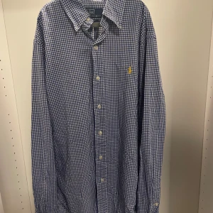 Rutig blå skjorta från Polo Ralph Lauren - Riktigt snygg blå och vit rutig skjorta från Polo Ralph Lauren i slim fit. Klassisk krage, långa ärmar och knäppning framtill. Ikoniska gula broderade loggan på bröstet. Perfekt för dig som gillar stilrena och tidlösa plagg.