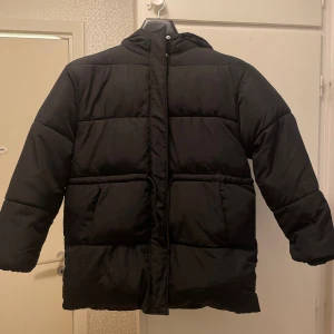 Svart pufferjacka med huva - Säljer en svart pufferjacka med huva och stora fickor framtill från H&M. Jackan är lång och stängs med dragkedja samt snören.