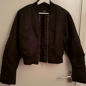 Svart bomberjacka från Gina Tricot - Säljer en svart bomberjacka från Gina Tricot med croppad passform. Jackan har en silvrig dragkedja framtill, ribbade muddar och klassisk bomberkrage. Perfekt att slänga på sig över en hoodie eller t-shirt för en cool look. Helt oanvänd