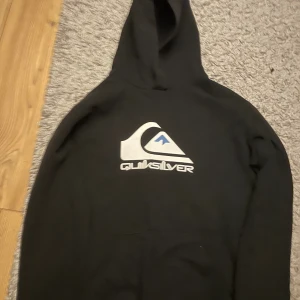 Svart Quiksilver hoodie med logga - Svart hoodie från Quiksilver med stor vit och blå logga på bröstet. Klassisk huva och magficka framtill. Perfekt för dig som gillar streetwear och vill ha en enkel men snygg tröja med tydlig branding.
