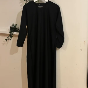 Svart långärmad abaya  - En stilren svart lång abaya med raka linjer och lång ärm. Klänningen har en enkel rund halsringning med liten slits framtill och är helt enfärgad utan mönster. Passformen är rak och loose, vilket ger en avslappnad och modern look.