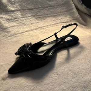 Svarta ballerinaskor Steve Madden Dazzled - Snygga svarta ballerinaskor från Steve Madden i lackat syntetmaterial. Skorna har spetsig tå, dekorativ rosett framtill och smala remmar med spänne runt hälen. Perfekta för dig som vill ha en stilren och trendig look.