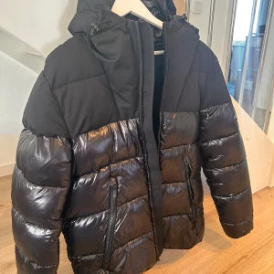 Svart pufferjacka från Replay - Snygg svart pufferjacka från Replay med glansiga partier och matta detaljer. Jackan har huva med tryck, två dragkedjefickor och markerad logga på ärmen. Perfekt för kalla dagar med sin rejäla vaddering och coola look.