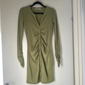 Olivgrön ribbad klänning från NA-KD - Olivgrön ribbad kortklänning från NA-KD med långärmad design och snygg rynkning framtill. Klänningen har knappar längs hela framsidan och är figurnära med stretchigt material som ger en slim passform. Perfekt för dig som gillar trendiga detaljer och stilren look.