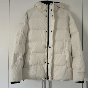 Vit pufferjacka med huva - Säljer en stilren vit pufferjacka med huva och svarta detaljer. Jackan har dragkedja och knappar framtill, två sidofickor och quiltad design. Perfekt för kalla dagar när du vill hålla värmen och ändå se fräsch ut.