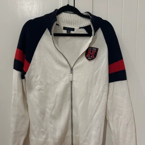 Vit kofta med dragkedja Tommy Hilfiger - Vit stickad kofta från Tommy Hilfiger med marinblå och röda detaljer på ärmarna. Dragkedja framtill och ribbad krage. Snyggt H-märke på bröstet och klassisk sportig look. Extremt liten i storlek, då de står Xl skulle jag mer säga storlek S.