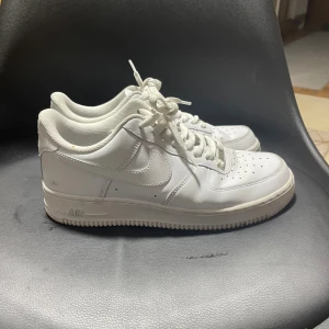 Nike Air Force 1 vita sneakers - Fräscha Nike Air Force 1 sneakers i helvitt skinn med klassisk rund tå och platt sula. Ikonisk design med perforerad tå, snörning och Nike-logga på sidan och hälen. Perfekt för dig som gillar cleana och tidlösa sneakers.