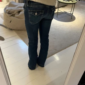 Blå bootcut jeans med rå kant - Snygga blå jeans med bootcut och rå, fransad kant nertill. Jeansen har markerade vita sömmar, fickor med lock och knapp bak samt klassisk femficksdesign. Materialet är jeans med stretch och de har låg midja för en avslappnad look.