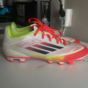Adidas fotbollsskor i vitt och rött - Säljer ett par Adidas fotbollsskor med snygg design i vitt, neon-gult och rött. Skorna har svarta ränder på sidorna, röda snören och dobbar för gräsplan. Lätta och smidiga med syntetmaterial och coola detaljer som sticker ut på planen.