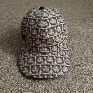 Ferragamo keps - Snygg keps från Salvatore Ferragamo med grått och svart mönster över hela kepsen. Justerbar rem baktill med metallspänne. Tillverkad i bomull och har en klassisk böjd skärm. Perfekt för dig som vill sticka ut med en exklusiv accessoar.