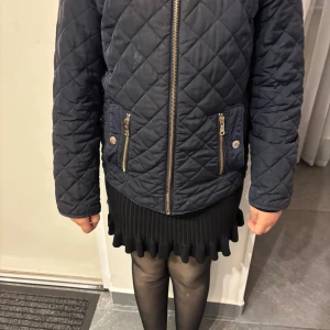 Mörkblå quiltad jacka från Zara - Quiltad mörkblå jacka från Zara med dragkedja framtill och två fickor med dragkedja och knappdetaljer. Jackan har lång ärm och snyggt rutmönster. Fodret är dekorerat med små vita stjärnor för en extra cool touch.