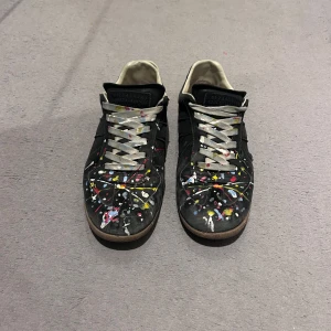 Maison Margiela sneakers med färgstänk - Maison Margiela sneakers i svart med unikt färgstänk i gult, vitt, blått och rött över tån och sidorna. Skorna har grå snörning, bruna gummisulor och klassisk låg profil. Perfekta för dig som vill sticka ut med en konstnärlig vibe.
