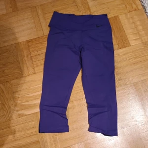 Lila Nike Dri-Fit korta leggings S - Snygga lila Nike Dri-Fit leggings i storlek S med svarta paneler längs sidorna. Modellen är trekvarts och har hög midja. Perfekta för träning med stretchigt och snabbtorkande material som andas.