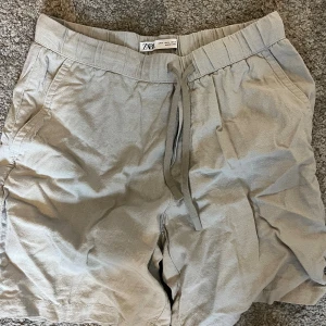 Beige shorts med snörning från Zara - Snygga beige shorts från Zara i storlek M. De har elastisk midja med snörning framtill, sidofickor och en bakficka med liten flik. Materialet är mjukt och luftigt, perfekt för varma dagar. Passformen är normal och stilen är enkel men fräsch.