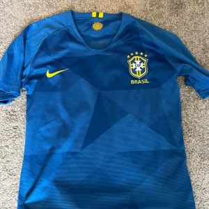 Blå Brasilien fotbollströja Nike - Säljer en officiell Brasilien fotbollströja från Nike i blått med geometriskt mönster. Tröjan har korta ärmar, gul Nike-logga och Brasilien-emblem med stjärnor på bröstet. Materialet är lätt och andas, perfekt för match eller träning.